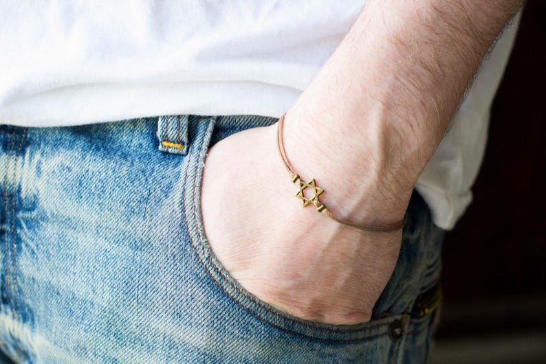 May include: A gold Star of David charm bracelet on a brown cord. The bracelet is worn on a person's wrist.