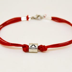 May include: Red string bracelet with a silver charm featuring the Libra zodiac symbol.