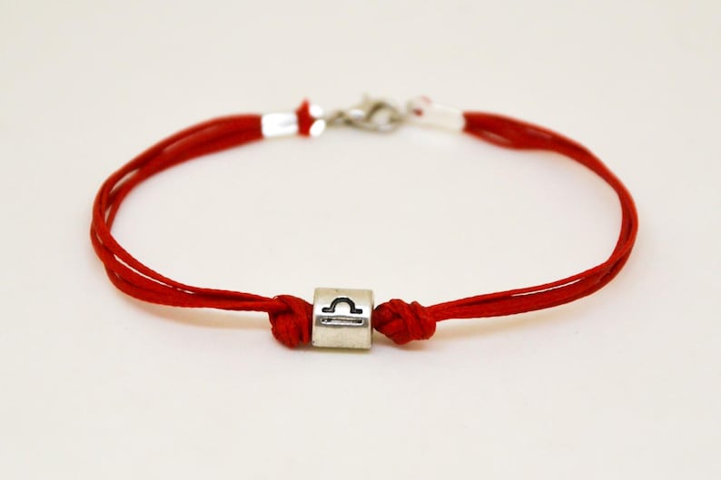 May include: Red string bracelet with a silver charm featuring the Libra zodiac symbol.