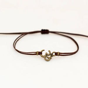May include: A brown string bracelet with an Om symbol charm. The charm is made of metal and has a gold finish.