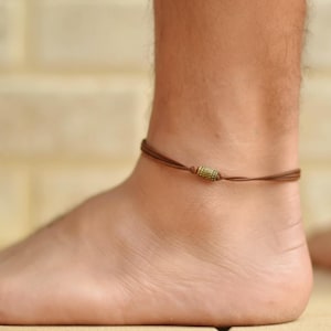 May include: A brown cord anklet featuring a single cylindrical brass-toned metal bead with a textured, ribbed surface. The anklet is constructed from thin, smooth brown cord material that wraps around the ankle with a simple, minimalist design.