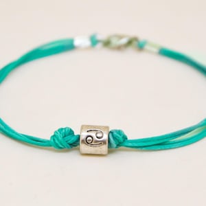May include: A turquoise blue string bracelet with a silver charm featuring the Cancer zodiac symbol.