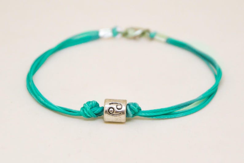 May include: A turquoise blue string bracelet with a silver charm featuring the Cancer zodiac symbol.