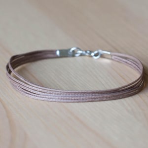 Könnte beinhalten: Ein zartes Armband mit mehreren Strängen aus hellbraunem Kordel. Das Armband wird mit einem silberfarbenen Verschluss gesichert. Das Design ist schlicht und elegant, geeignet für den täglichen Gebrauch. Das Armband wird auf einer hellen Holzoberfläche gezeigt.