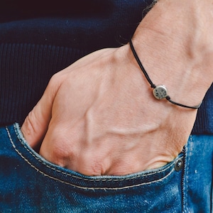 Op de afbeelding: Een close-up van iemands pols met een zwart koordarmband met een ronde zilveren hanger. De hand van de persoon zit in een zak van een blauwe spijkerbroek.