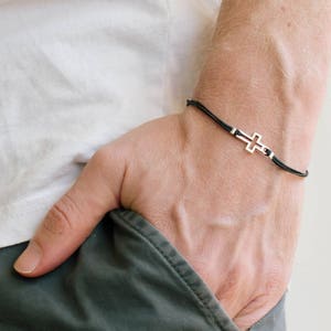 Könnte beinhalten: Ein Armband aus schwarzem Band mit einem silbernen Kreuzanhänger.