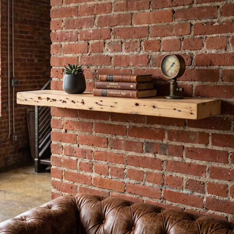May include: A thick, light-colored wooden floating shelf mounted on a red brick wall. The shelf holds a small potted succulent, a stack of three books titled Urban Artifacts and Industrial Age, and a round metal pressure gauge on a stand.