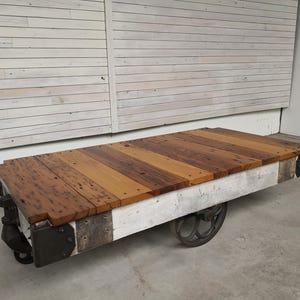 May include: A rustic industrial coffee table, repurposed from a vintage industrial cart. The top is made of reclaimed wood planks in shades of brown and tan, arranged in a striped pattern. The base is painted white with visible metal accents and features two sturdy wheels. This unique piece blends rustic charm with industrial style, perfect for adding character to any living space.