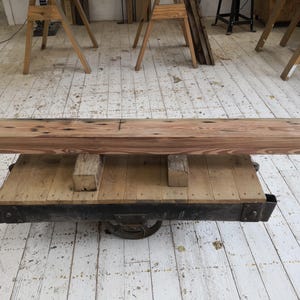 K&ouml;nnte beinhalten: Ein rechteckiger, hellbrauner Holzbalken mit dunklen Knoten liegt auf einer Holzplattform mit einem schwarzen Metallrahmen. Die Plattform wird von einem Metallrad getragen. Der Balken ist wahrscheinlich Teil eines rustikalen M&ouml;belst&uuml;cks.