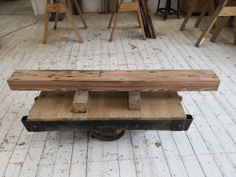 K&ouml;nnte beinhalten: Ein rechteckiger, hellbrauner Holzbalken mit dunklen Knoten liegt auf einer Holzplattform mit einem schwarzen Metallrahmen. Die Plattform wird von einem Metallrad getragen. Der Balken ist wahrscheinlich Teil eines rustikalen M&ouml;belst&uuml;cks.