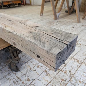 May include: A weathered, rectangular wooden beam with visible grain and cracks. The beam is light gray with darker gray accents and is supported by a metal cart with wheels. The wood appears to be reclaimed or rustic.