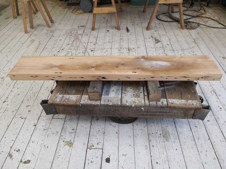 Pu&ograve; includere: Un lungo ripiano in legno rustico realizzato con legno riciclato, con nodi e imperfezioni, che poggia su un carrello di metallo con ruote. Il ripiano &egrave; di colore marrone chiaro e ha una finitura naturale e invecchiata.