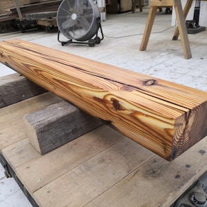 K&ouml;nnte beinhalten: Ein gro&szlig;er, rechteckiger Holzbalken mit nat&uuml;rlicher, goldbrauner Oberfl&auml;che. Der Balken, etwa 152 cm lang, ruht auf zwei kleineren Holzbl&ouml;cken. Die Holzmaserung ist sichtbar, und ein Riss verl&auml;uft entlang der Oberseite. Der Balken ist wahrscheinlich f&uuml;r den Bau oder die Dekoration.