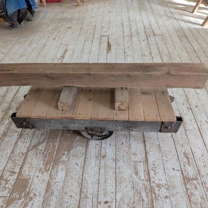 K&ouml;nnte beinhalten: Ein rustikaler Holzbalken liegt auf einem umfunktionierten Industriefahrwagen. Der Balken ist hellbraun mit sichtbarer Holzmaserung. Der Wagen hat eine Holzplattform und einen Metallrahmen mit R&auml;dern, was eine einzigartige, Vintage-&Auml;sthetik erzeugt.