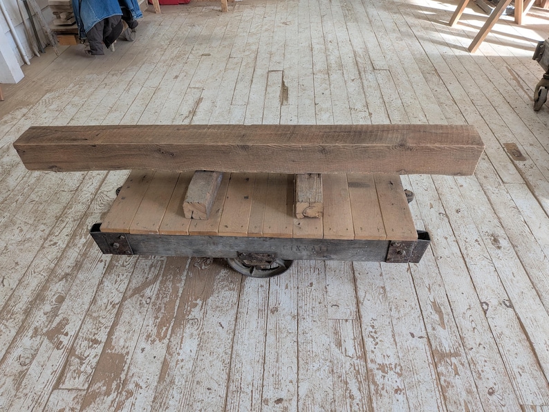 K&ouml;nnte beinhalten: Ein rustikaler Holzbalken liegt auf einem umfunktionierten Industriefahrwagen. Der Balken ist hellbraun mit sichtbarer Holzmaserung. Der Wagen hat eine Holzplattform und einen Metallrahmen mit R&auml;dern, was eine einzigartige, Vintage-&Auml;sthetik erzeugt.