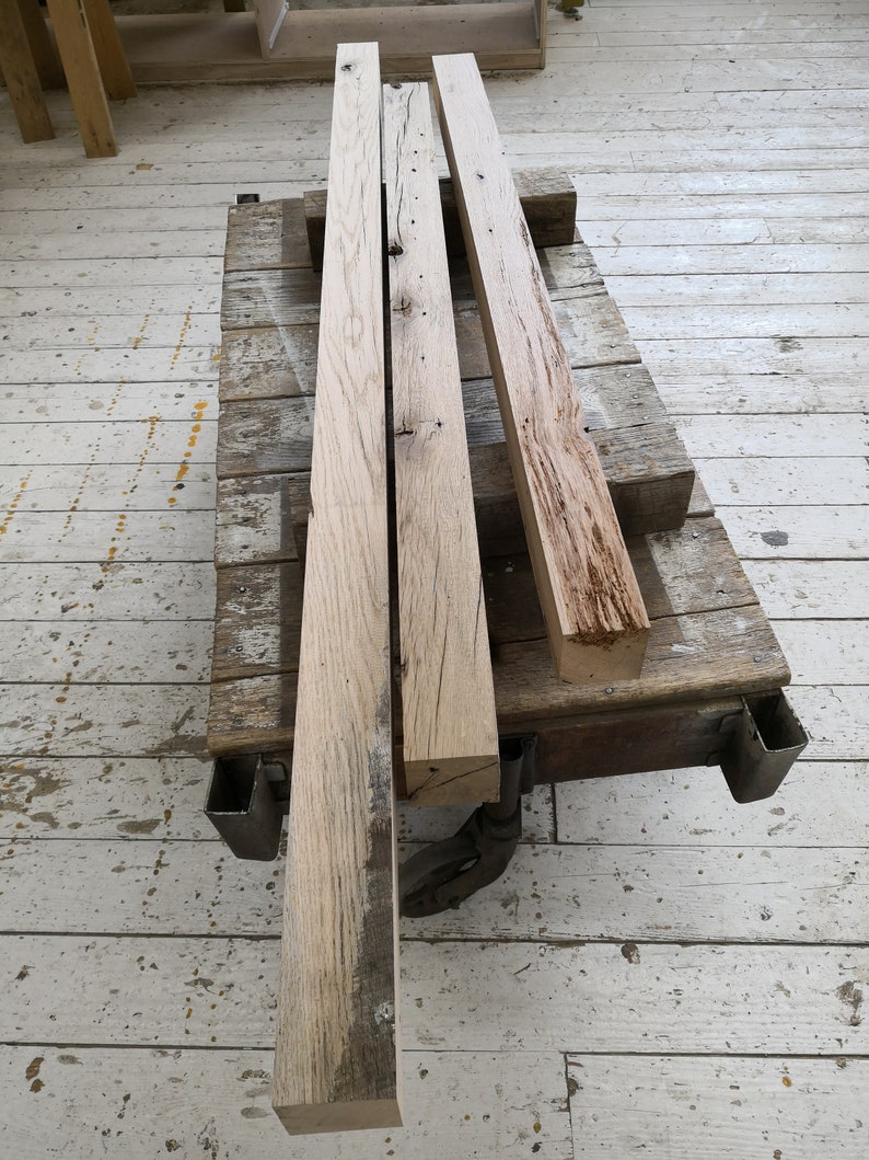 Puede incluir: Tres piezas de tablones de madera toscamente labrados y desgastados en un carro de madera. Los tablones son de color marr&oacute;n claro con nudos y patrones de veta de color marr&oacute;n oscuro.