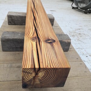 K&ouml;nnte beinhalten: Ein rechteckiger, verwitterter Holzbalken mit einem deutlichen Riss, der sich &uuml;ber seine gesamte L&auml;nge erstreckt. Das Holz hat einen warmen, goldbraunen Farbton mit sichtbaren Maserungen und Knoten. Der Balken liegt auf zwei kleineren, grauen Holzbl&ouml;cken.