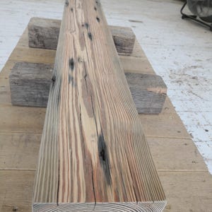 K&ouml;nnte beinhalten: Ein verwitterter, rechteckiger Holzbalken mit sichtbarer Maserung und &Auml;sten. Der Balken ist hellbraun mit dunkleren Streifen und kreisf&ouml;rmigen Jahresringen an einem Ende. Er liegt auf zwei kleineren Holzbl&ouml;cken.
