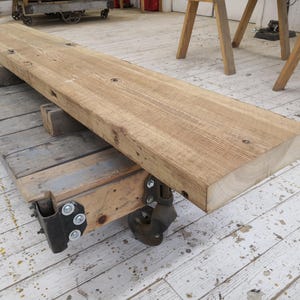 May include: A long, rustic wooden plank on a wooden dolly with wheels. The plank is light brown and has a rough, weathered surface. The dolly is made of wood and metal and has four wheels.