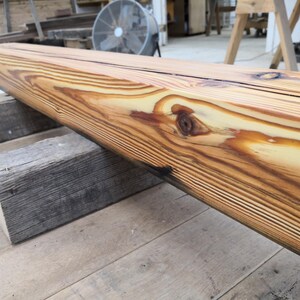 K&ouml;nnte beinhalten: Ein gro&szlig;er, unfertiger Holzbalken mit einem nat&uuml;rlichen Holzmaserungsmuster. Der Balken wird von zwei verwitterten, grauen Holzbl&ouml;cken getragen. Das Holz hat eine warme, goldbraune Farbe mit dunkleren Streifen und Knoten, was darauf hindeutet, dass es aus Kiefernholz besteht.