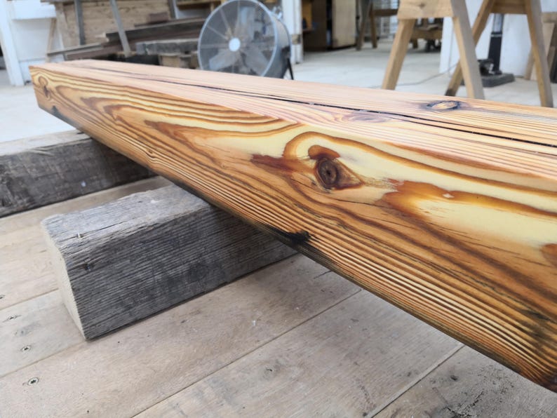 K&ouml;nnte beinhalten: Ein gro&szlig;er, unfertiger Holzbalken mit einem nat&uuml;rlichen Holzmaserungsmuster. Der Balken wird von zwei verwitterten, grauen Holzbl&ouml;cken getragen. Das Holz hat eine warme, goldbraune Farbe mit dunkleren Streifen und Knoten, was darauf hindeutet, dass es aus Kiefernholz besteht.