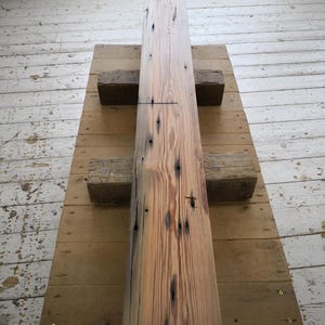 K&ouml;nnte beinhalten: Ein langer, rechteckiger Holzbalken mit nat&uuml;rlichem, verwittertem Aussehen. Der Balken ist hellbraun mit dunkleren Knoten und Unvollkommenheiten. Er ruht auf einer Holzplattform, die von zwei kleineren Holzbl&ouml;cken getragen wird. Die Holzmaserung ist sichtbar.
