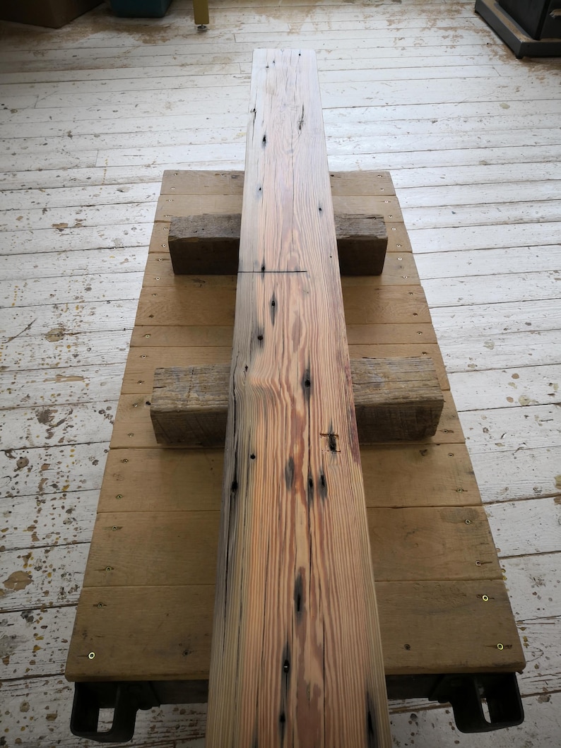 K&ouml;nnte beinhalten: Ein langer, rechteckiger Holzbalken mit nat&uuml;rlichem, verwittertem Aussehen. Der Balken ist hellbraun mit dunkleren Knoten und Unvollkommenheiten. Er ruht auf einer Holzplattform, die von zwei kleineren Holzbl&ouml;cken getragen wird. Die Holzmaserung ist sichtbar.