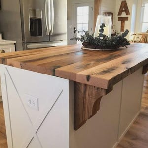 K&ouml;nnte beinhalten: Rustikale K&uuml;cheninsel mit einer dicken Arbeitsplatte aus Altholz. Der Sockel der Insel ist wei&szlig; gestrichen und hat ein X-f&ouml;rmiges Design. Eine dekorative Holzhalterung st&uuml;tzt die Arbeitsplatte. Im Hintergrund ist ein K&uuml;hlschrank aus Edelstahl zu sehen.
