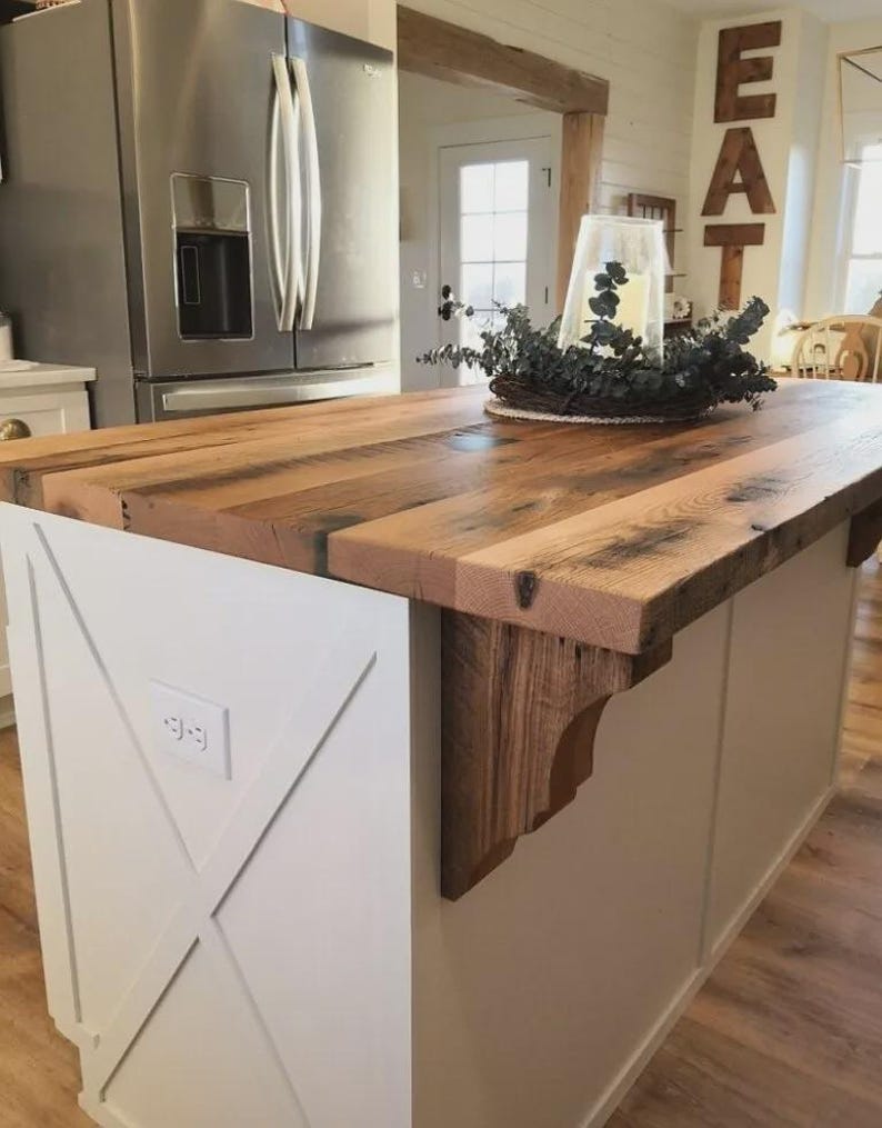 K&ouml;nnte beinhalten: Rustikale K&uuml;cheninsel mit einer dicken Arbeitsplatte aus Altholz. Der Sockel der Insel ist wei&szlig; gestrichen und hat ein X-f&ouml;rmiges Design. Eine dekorative Holzhalterung st&uuml;tzt die Arbeitsplatte. Im Hintergrund ist ein K&uuml;hlschrank aus Edelstahl zu sehen.