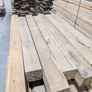 K&ouml;nnte beinhalten: Ein Stapel grob behauener, rechteckiger Holzbalken in nat&uuml;rlicher, hellbrauner Farbe. Die Balken weisen sichtbare Holzmaserung und Knoten auf, was auf eine rustikale oder recycelte &Auml;sthetik hindeutet. Weitere Holzstapel sind im Hintergrund zu sehen.