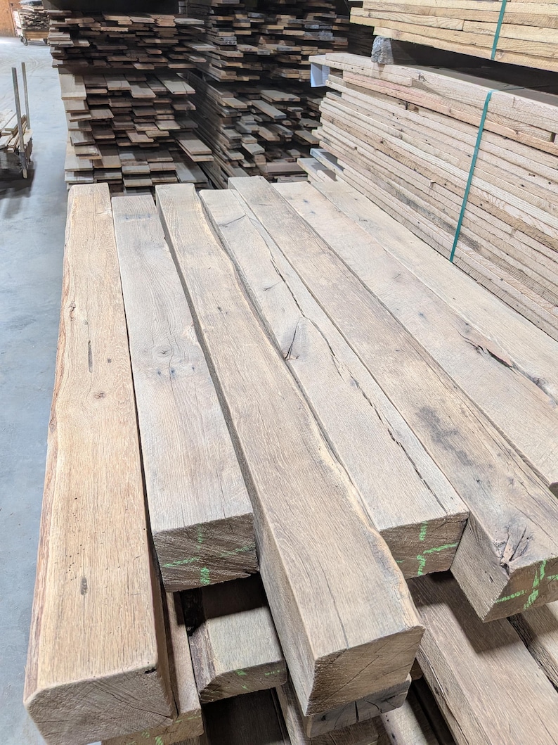 K&ouml;nnte beinhalten: Ein Stapel grob behauener, rechteckiger Holzbalken in nat&uuml;rlicher, hellbrauner Farbe. Die Balken weisen sichtbare Holzmaserung und Knoten auf, was auf eine rustikale oder recycelte &Auml;sthetik hindeutet. Weitere Holzstapel sind im Hintergrund zu sehen.