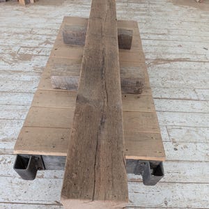 K&ouml;nnte beinhalten: Ein verwitterter, rechteckiger Holzbalken mit rauer, strukturierter Oberfl&auml;che. Der Balken hat eine nat&uuml;rliche braune Farbe und scheint aus Altholz zu bestehen. Er liegt auf einer Holzplattform mit Metallst&uuml;tzen, was auf eine rustikale oder industrielle &Auml;sthetik hindeutet. Abmessungen: 20 cm x 20 cm x 244 cm.
