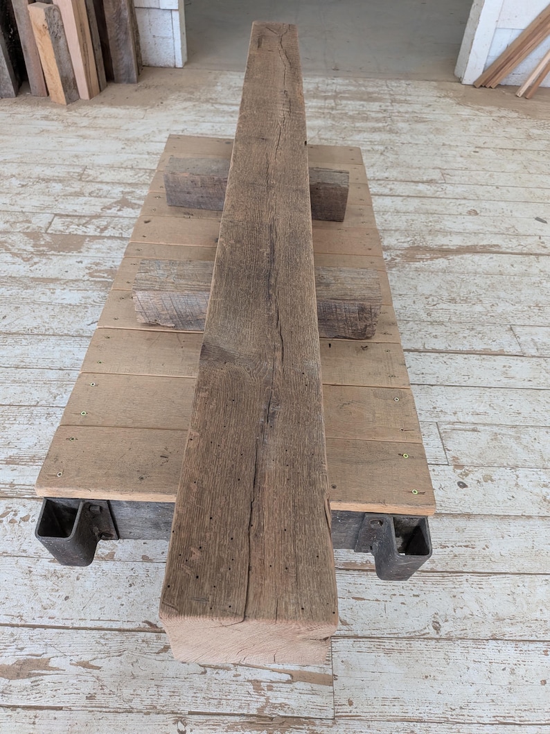 K&ouml;nnte beinhalten: Ein verwitterter, rechteckiger Holzbalken mit rauer, strukturierter Oberfl&auml;che. Der Balken hat eine nat&uuml;rliche braune Farbe und scheint aus Altholz zu bestehen. Er liegt auf einer Holzplattform mit Metallst&uuml;tzen, was auf eine rustikale oder industrielle &Auml;sthetik hindeutet. Abmessungen: 20 cm x 20 cm x 244 cm.