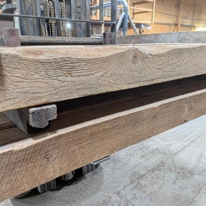 K&ouml;nnte beinhalten: Nahaufnahme von gestapelten, verwitterten Holzbalken. Die Balken haben eine nat&uuml;rliche, hellbraune Farbe mit sichtbarer Holzmaserung und Knoten. Das Holz scheint recycelt oder rustikal zu sein, mit einer rauen Textur. Die Balken sind auf einer Palette gestapelt.