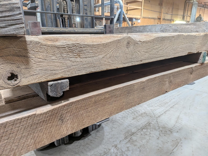 K&ouml;nnte beinhalten: Nahaufnahme von gestapelten, verwitterten Holzbalken. Die Balken haben eine nat&uuml;rliche, hellbraune Farbe mit sichtbarer Holzmaserung und Knoten. Das Holz scheint recycelt oder rustikal zu sein, mit einer rauen Textur. Die Balken sind auf einer Palette gestapelt.