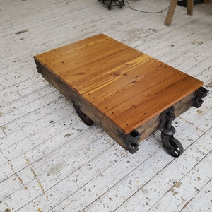May include: A wooden coffee table with a rustic brown finish. The table is made from reclaimed wood and has a metal frame. The table has four wheels and is sitting on a white wooden floor.