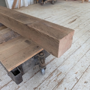 May include: A large, weathered, rectangular wooden beam with a rough, textured surface. The beam is a natural brown color and rests on a wooden cart with metal wheels. The cart has a wooden top and is also made of weathered wood.