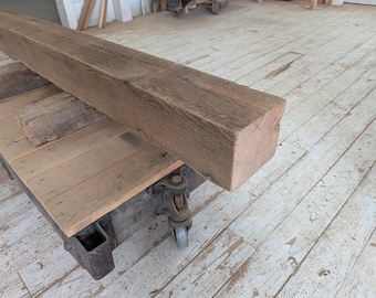 May include: A large, weathered, rectangular wooden beam with a rough, textured surface. The beam is a natural brown color and rests on a wooden cart with metal wheels. The cart has a wooden top and is also made of weathered wood.