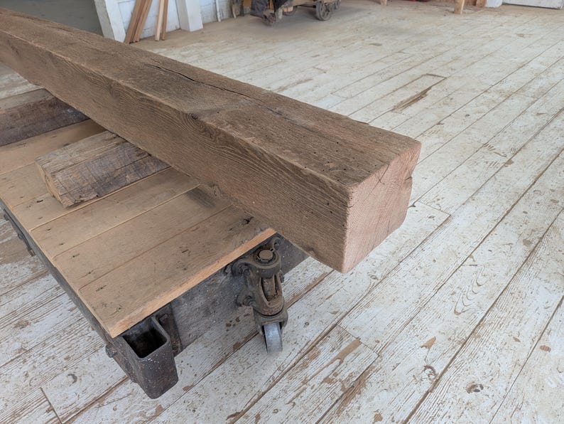 K&ouml;nnte beinhalten: Ein gro&szlig;er, verwitterter, rechteckiger Holzbalken mit rauer, strukturierter Oberfl&auml;che. Der Balken hat eine nat&uuml;rliche braune Farbe und liegt auf einem Holzwagen mit Metallr&auml;dern. Der Wagen hat eine Holzplatte und ist ebenfalls aus verwittertem Holz gefertigt.