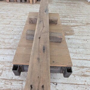 May include: A weathered, rectangular wooden beam with visible grain and knots, resting on a wooden platform supported by metal brackets. Two smaller wooden blocks are placed on the platform. The wood has a natural, aged appearance.