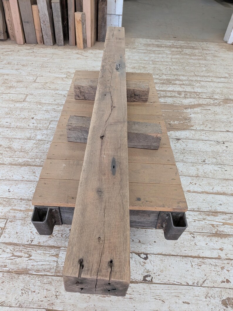 May include: A weathered, rectangular wooden beam with visible grain and knots, resting on a wooden platform supported by metal brackets. Two smaller wooden blocks are placed on the platform. The wood has a natural, aged appearance.