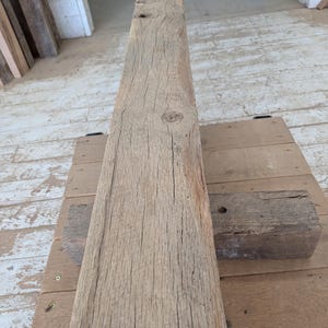 K&ouml;nnte beinhalten: Ein verwitterter, rechteckiger Holzbalken in nat&uuml;rlicher, hellbrauner Farbe. Die Holzmaserung ist sichtbar, mit zahlreichen Rissen und Knoten. Der Balken liegt auf einer Holzoberfl&auml;che, was auf ein rustikales oder recyceltes Holzprodukt hindeutet.