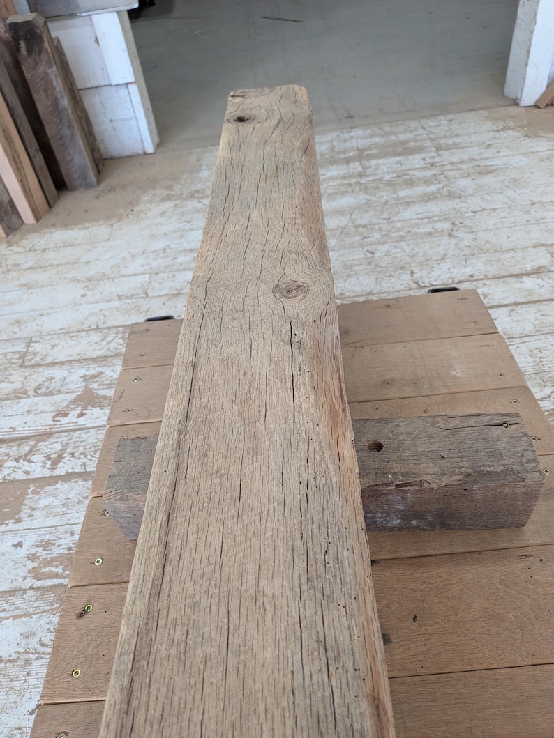 K&ouml;nnte beinhalten: Ein verwitterter, rechteckiger Holzbalken in nat&uuml;rlicher, hellbrauner Farbe. Die Holzmaserung ist sichtbar, mit zahlreichen Rissen und Knoten. Der Balken liegt auf einer Holzoberfl&auml;che, was auf ein rustikales oder recyceltes Holzprodukt hindeutet.