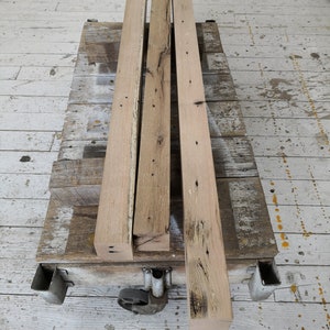 Puede incluir: Tres piezas de madera recuperada, cada una con un patr&oacute;n de veta diferente y nudos, est&aacute;n apiladas en un carro de metal. La madera es de color marr&oacute;n claro y tiene un aspecto r&uacute;stico.