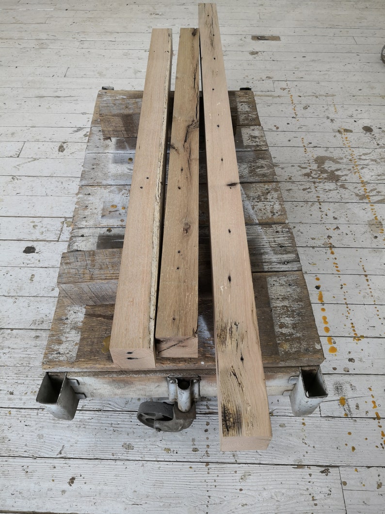 Puede incluir: Tres piezas de madera recuperada, cada una con un patr&oacute;n de veta diferente y nudos, est&aacute;n apiladas en un carro de metal. La madera es de color marr&oacute;n claro y tiene un aspecto r&uacute;stico.