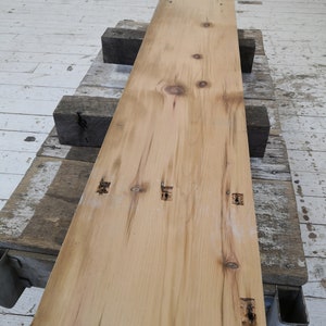 Pu&ograve; includere: Una lunga tavola di legno marrone chiaro con nodi e fori per chiodi. La tavola &egrave; appoggiata su altre tavole di legno.