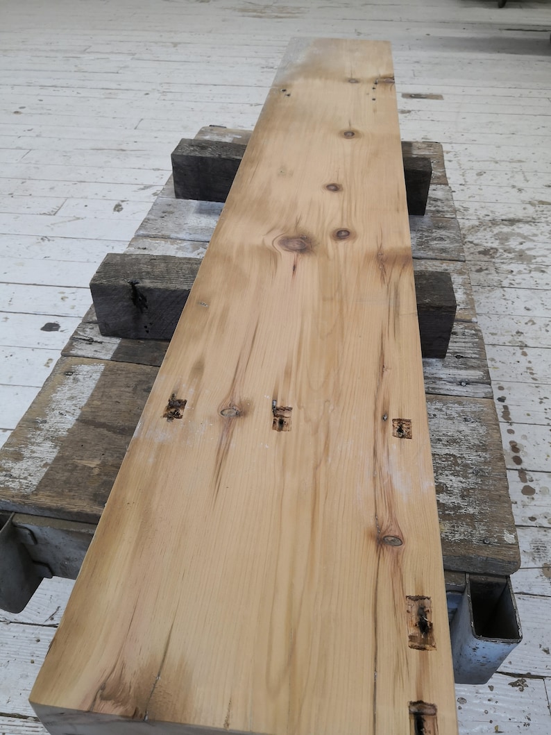 Pu&ograve; includere: Una lunga tavola di legno marrone chiaro con nodi e fori per chiodi. La tavola &egrave; appoggiata su altre tavole di legno.
