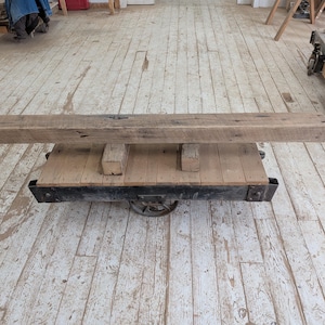 May include: A rustic, rectangular wooden beam rests on a vintage cart with a black metal frame and wheels. The beam is a natural, weathered brown color, and the cart has a wooden platform.