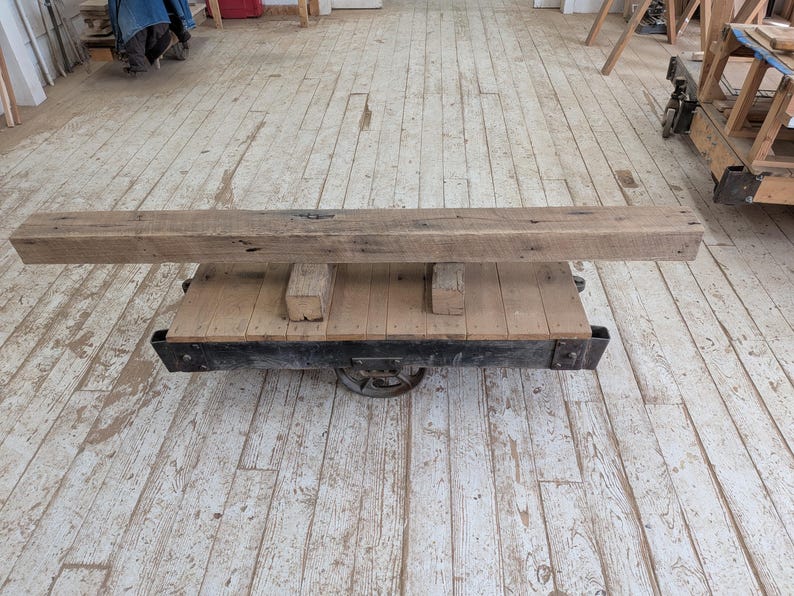 May include: A rustic, rectangular wooden beam rests on a vintage cart with a black metal frame and wheels. The beam is a natural, weathered brown color, and the cart has a wooden platform.
