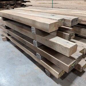 May include: A stack of rough-hewn, rectangular wooden beams. The beams are a natural, light brown color with visible wood grain and some green markings. They are stacked horizontally, supported by smaller wooden blocks.